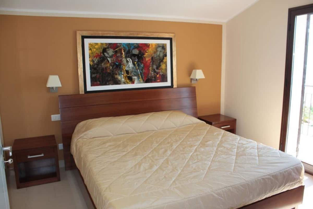 Une chambre à coucher avec un grand lit, une tête de lit en bois, deux tables de chevet avec des lampes et un tableau abstrait aux couleurs vives accroché au mur.