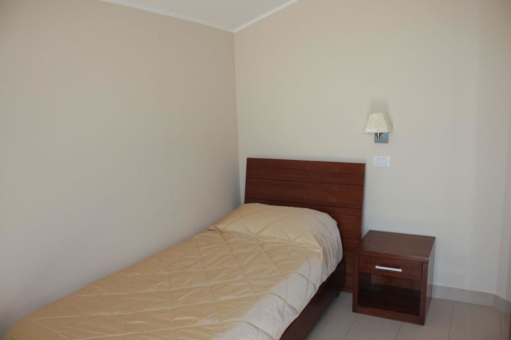 Una cama individual con cabecera de madera en una habitación con paredes lisas y una mesita de noche pequeña con una lámpara.