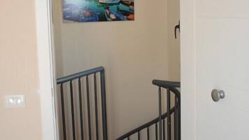 Un pasillo con un cuadro de una escena costera en la pared, una escalera con barandilla negra y una puerta blanca.