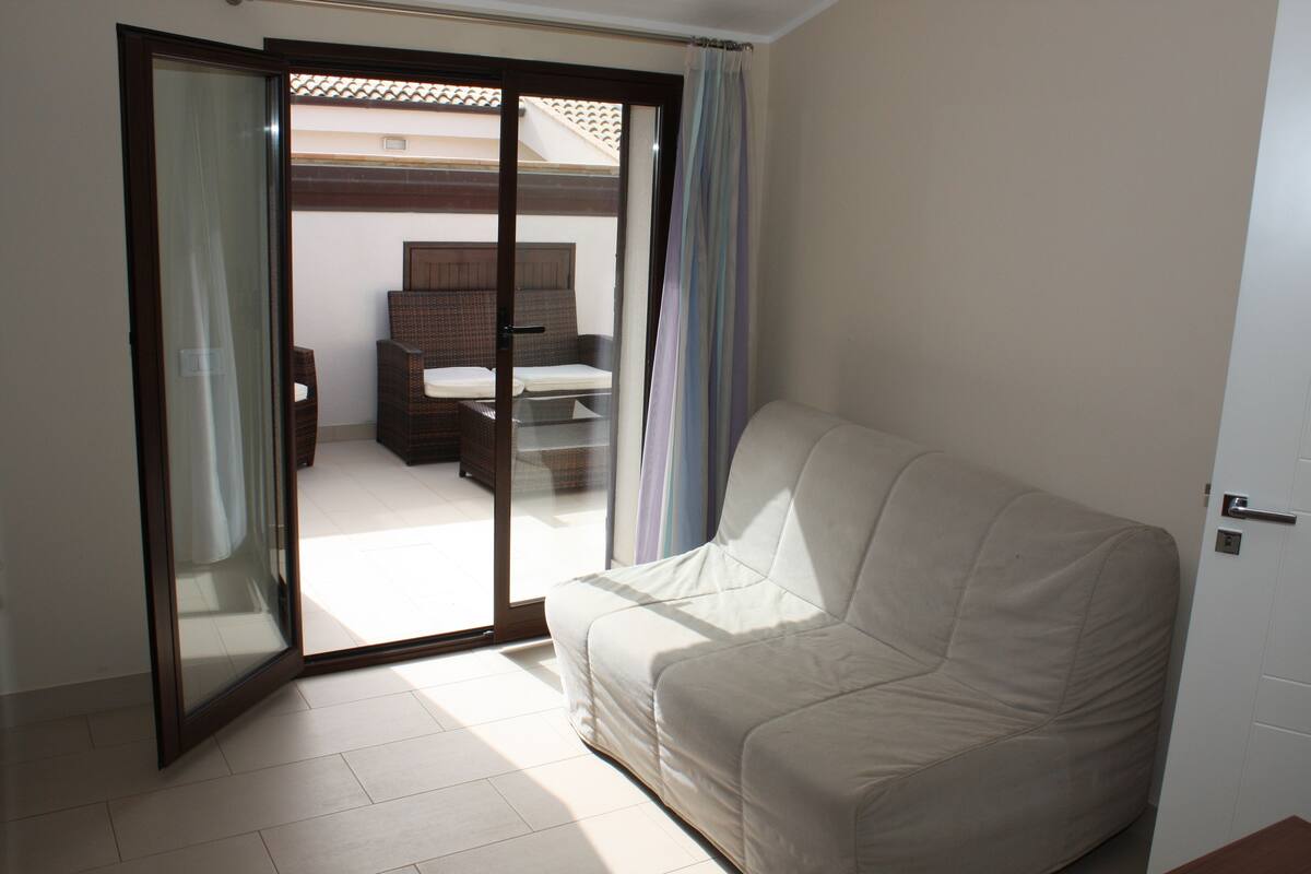 Une pièce dotée d’une porte-fenêtre coulissante donnant sur un balcon, d’un canapé blanc et d’un lit avec une tête de lit.