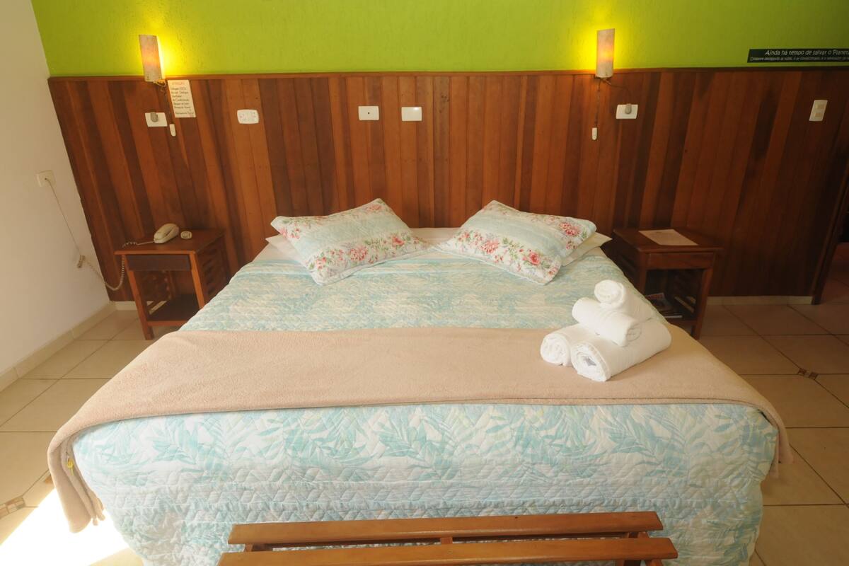 A hotel room with a wooden headboard, a bed with floral bedding, a bedside table with a phone, and a bench at the foot of the bed.