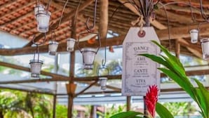 Een buitenterras met houten tafels en stoelen, lantaarns die aan het plafond hangen, en een label met de tekst 'DANCE AME CANTE VIVA'.