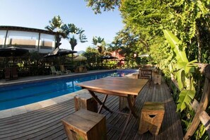 Une terrasse en bois avec une piscine, entourée d’une végétation luxuriante et ombragée par des parasols.