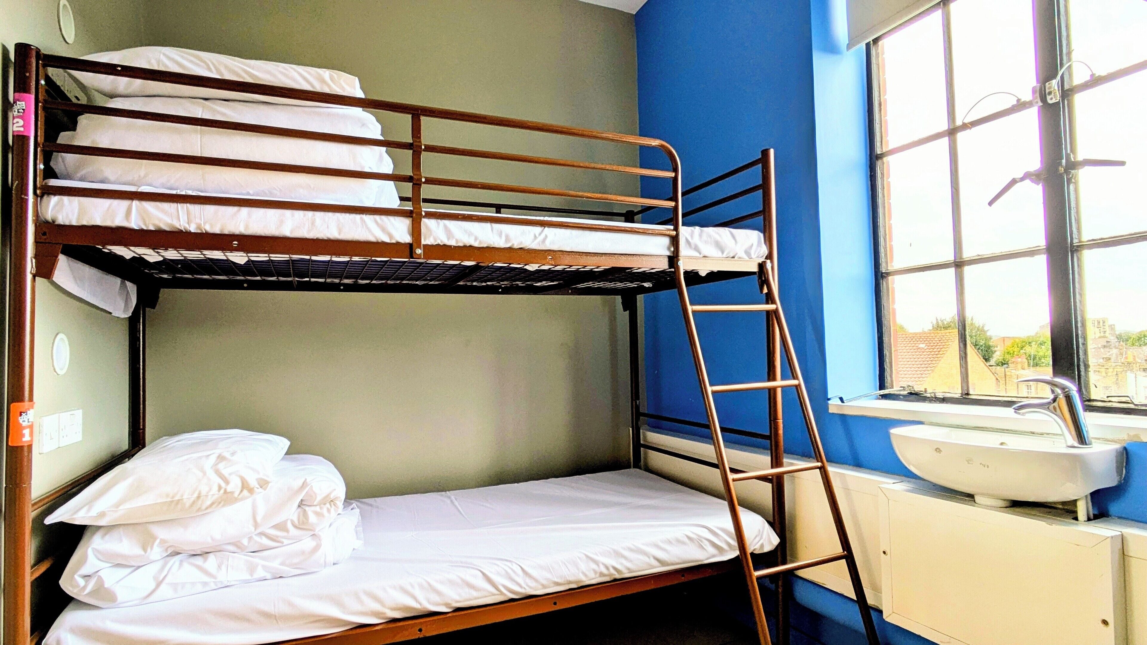 A bunk bed room with a ladder, a sink, and a window with a view of buildings.