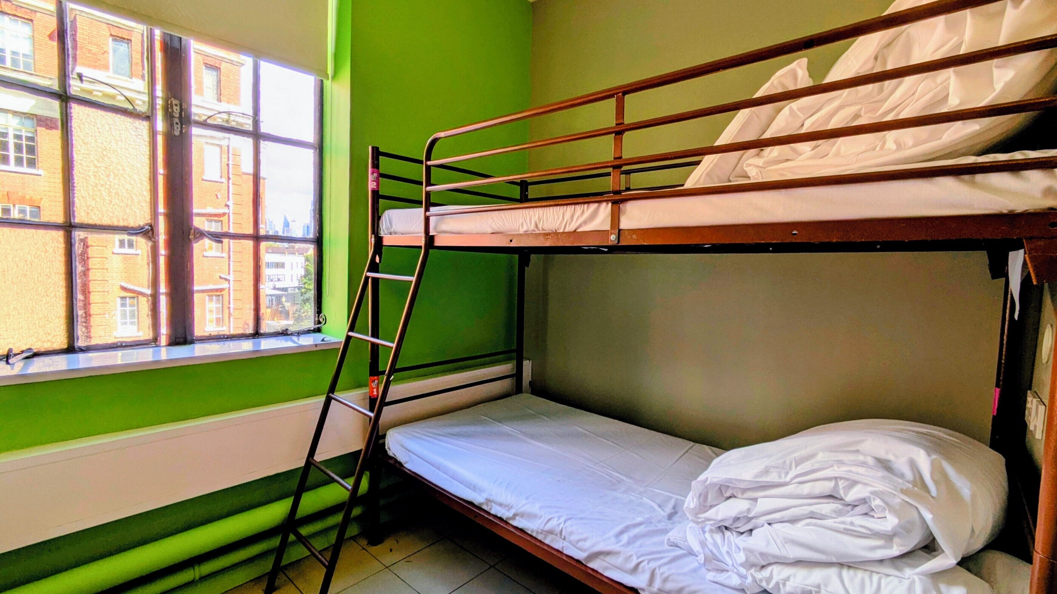 Een slaapkamer met stapelbed, raam en uitzicht op gebouwen.
