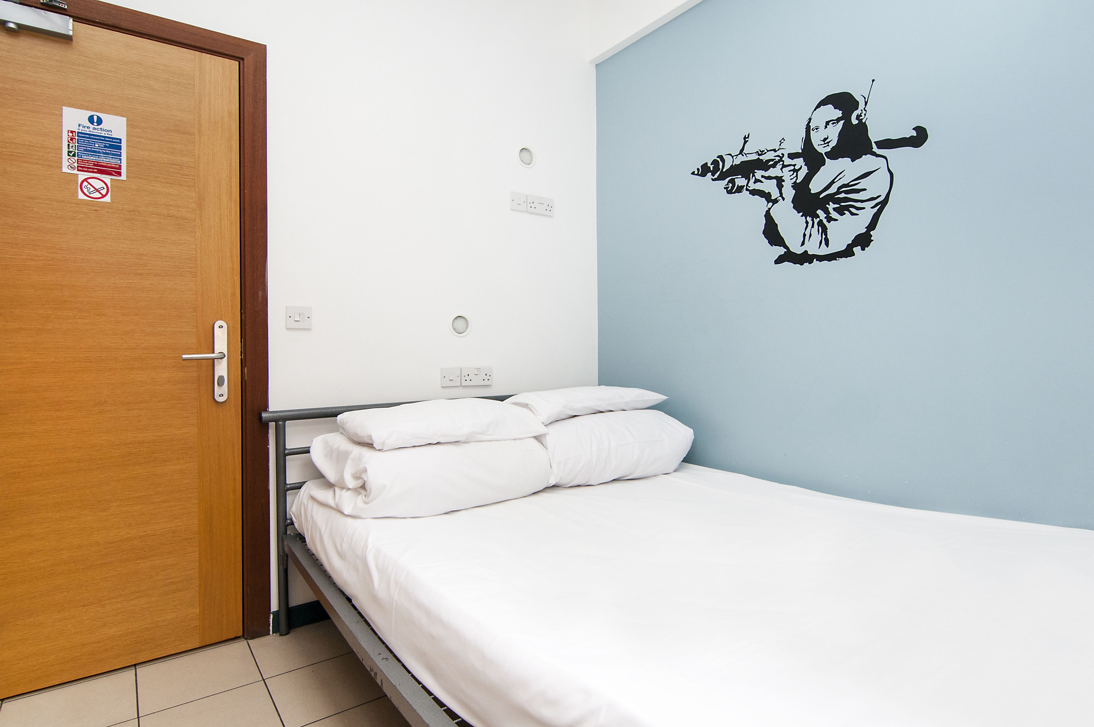 A bedroom with a wooden door, a bed with white bedding, a wall decal of a person playing a flute, and a sign on the door.
