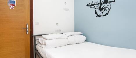 A bedroom with a wooden door, a bed with white bedding, a wall decal of a person playing a flute, and a sign on the door.