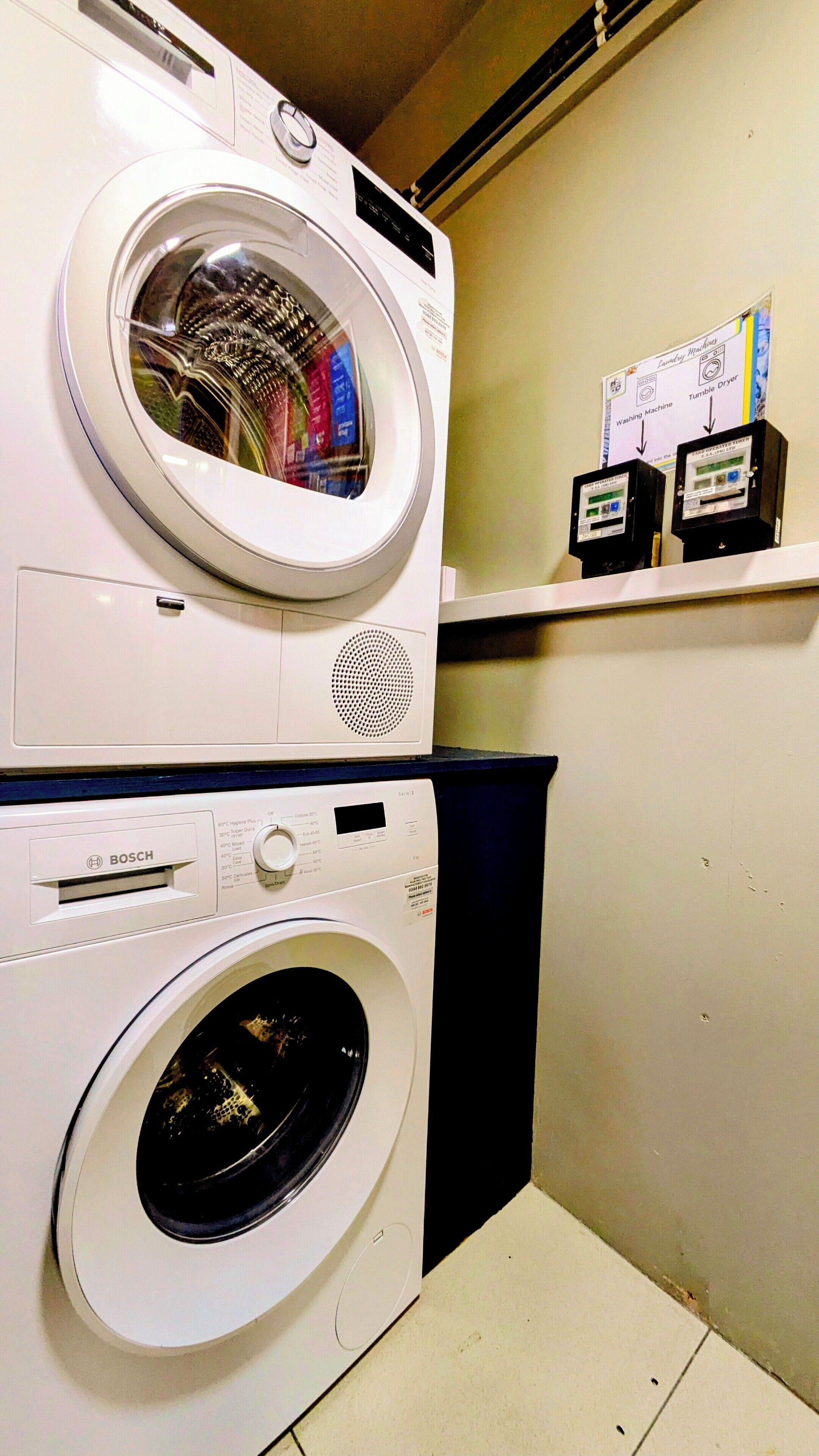 Two stacked front-loading washing machines in a laundry room with a shelf above displaying a diagram of laundry machines.