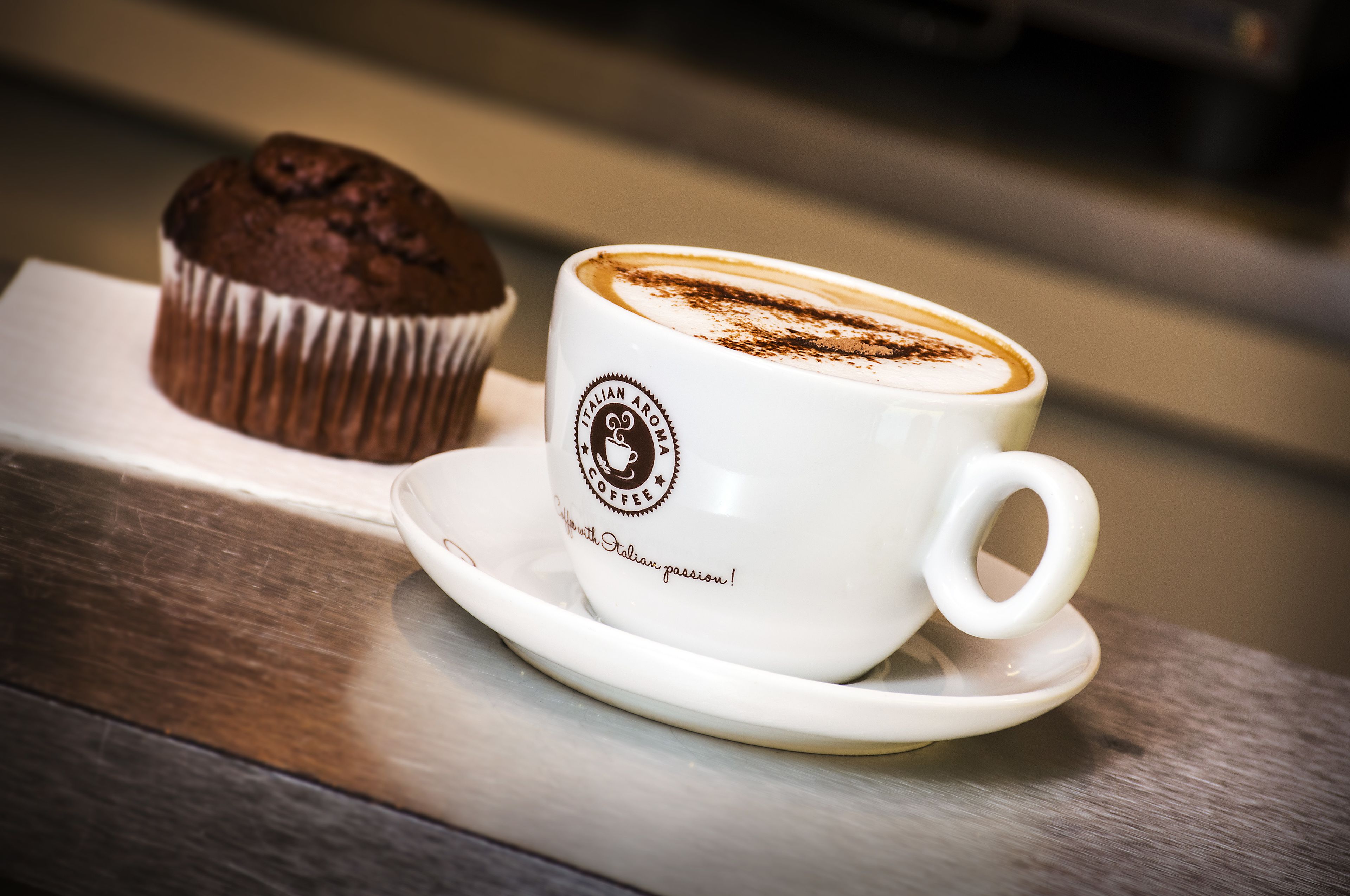 A white cup with a logo and text, a muffin, and a saucer on a wooden surface.