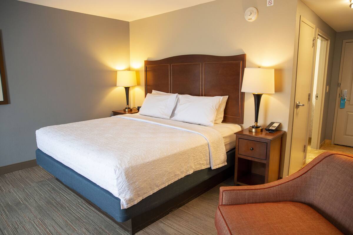 A hotel room with a large bed, two bedside tables with lamps, a chair, and a door.