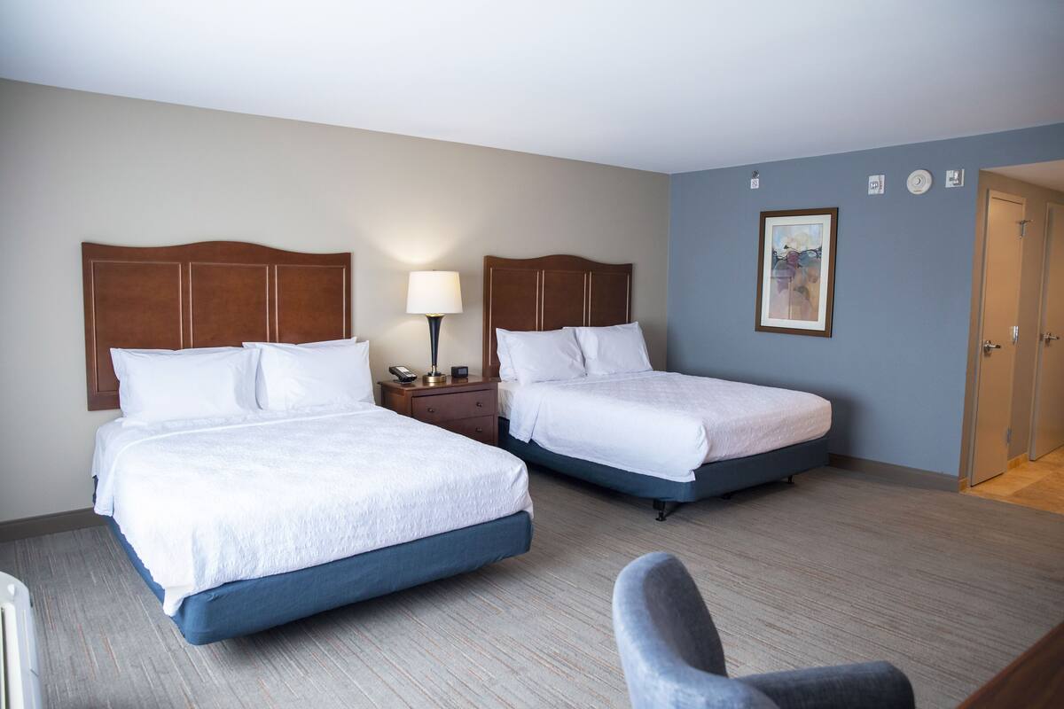 A hotel room with two beds, a nightstand with a lamp, a framed picture on the wall, and a door.