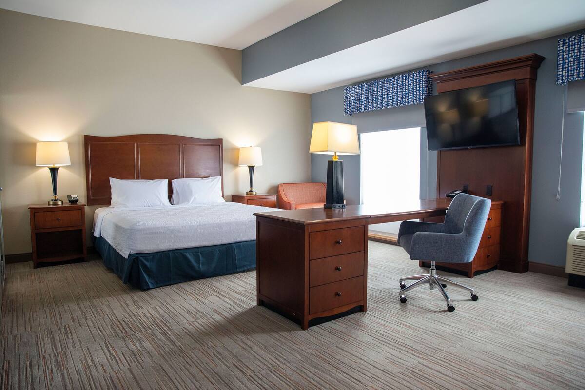 A hotel room with a bed, desk, chair, lamps, and a TV.