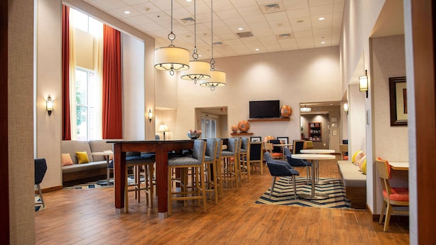 A spacious hotel lobby with a wooden floor, a bar area with stools, a seating area with sofas, and a television mounted on the wall.