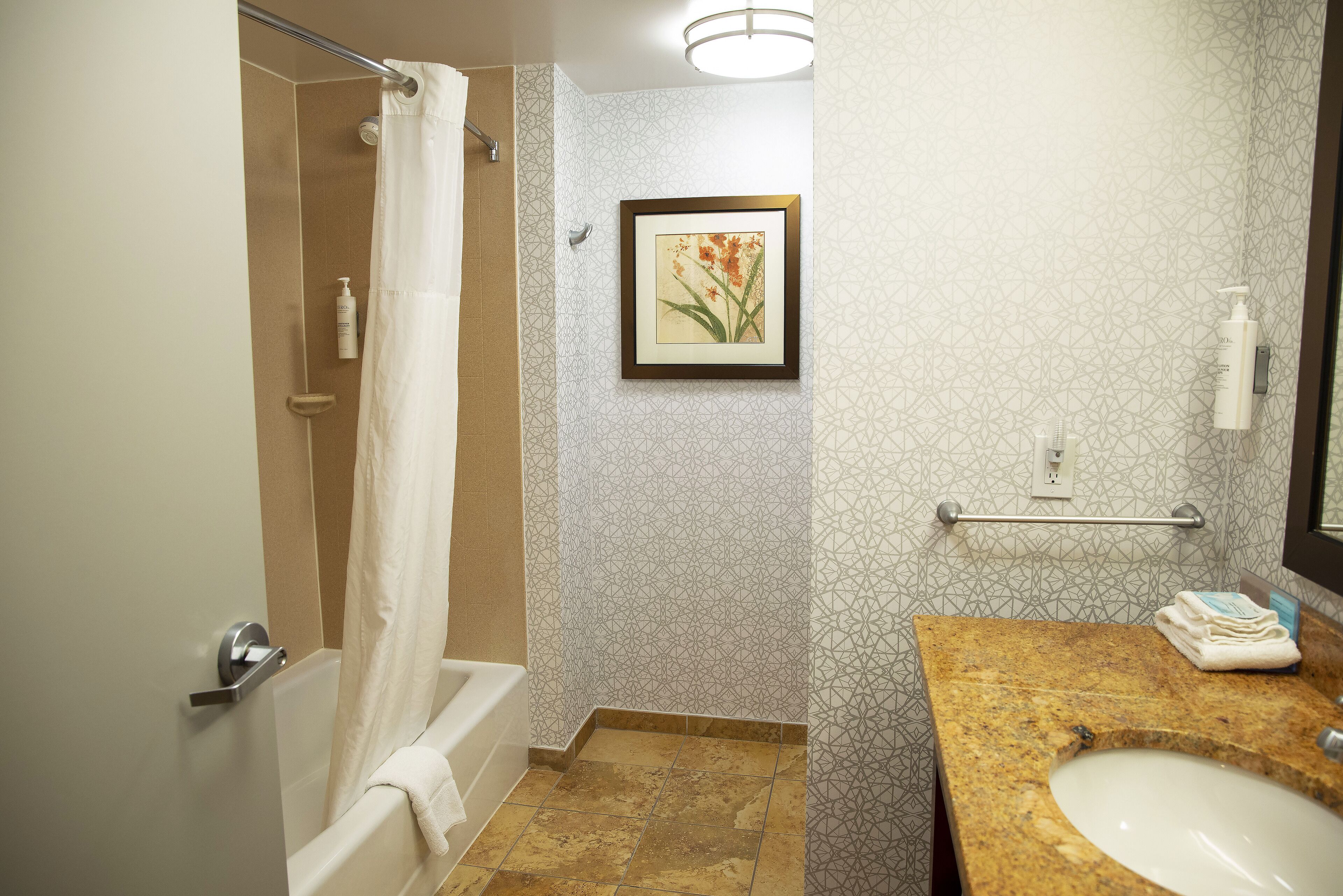 A bathroom with a shower, a framed picture on the wall, a sink with toiletries, and a towel rack.