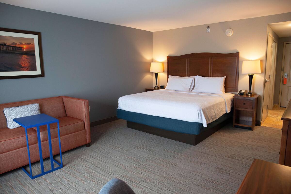 A hotel room with a bed, a nightstand, a bedside table, a sofa, and a framed picture on the wall.