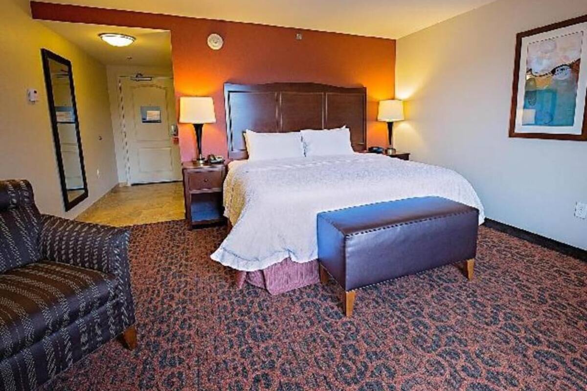 A hotel room with a bed, bedside lamps, a bench, a chair, and a painting on the wall.