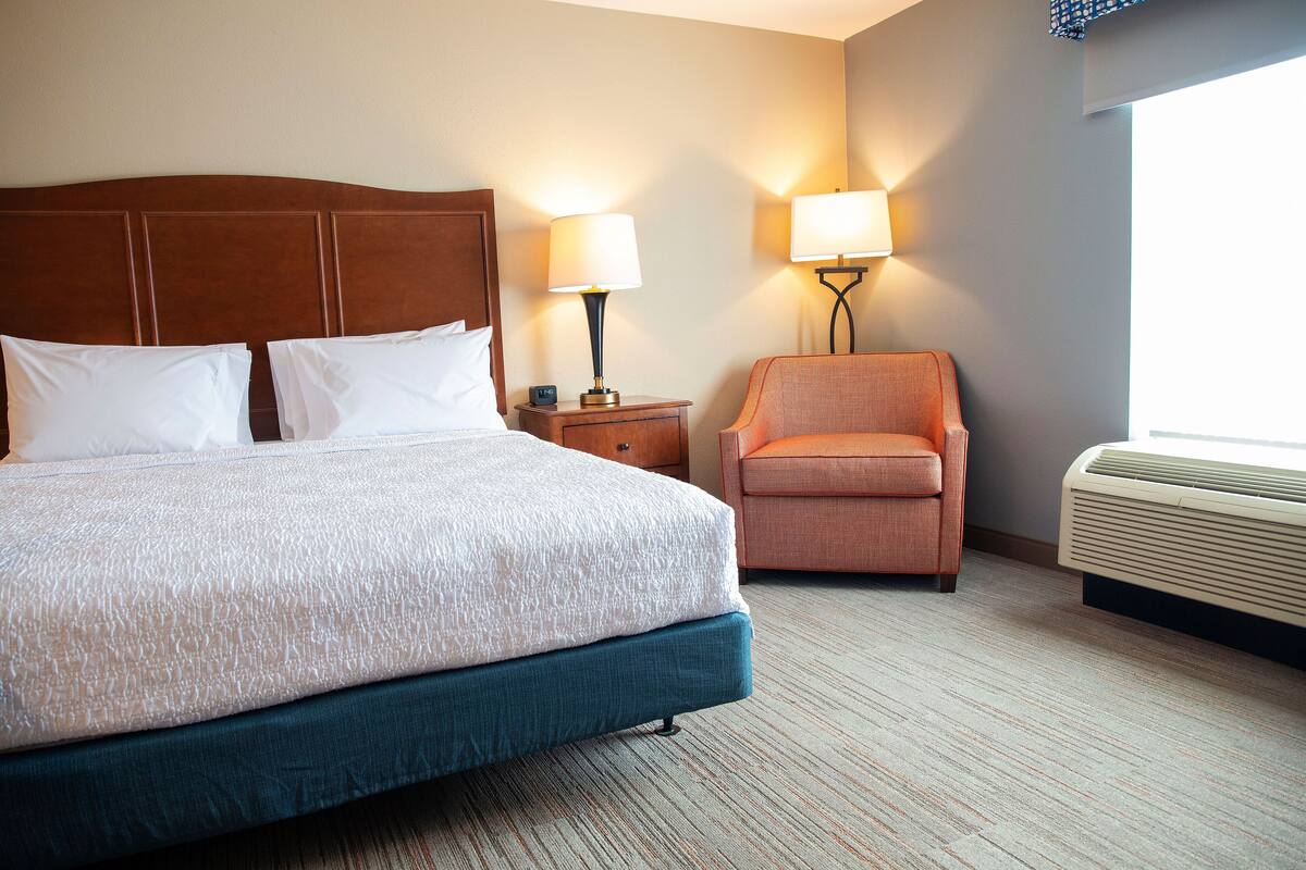 A hotel room with a large bed, a nightstand with a lamp, a chair, and a wall-mounted air conditioner.