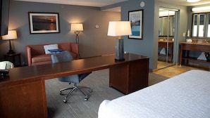 A hotel room with a bed, a desk, a chair, a sofa, a lamp, a painting, and a bathroom with a mirror and sink.