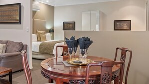 A dining area with a round wooden table, chairs, and a tray with glasses and napkins. A bed with a beige headboard and pillows is visible in the background. - 53 on Victoria (Warwick)