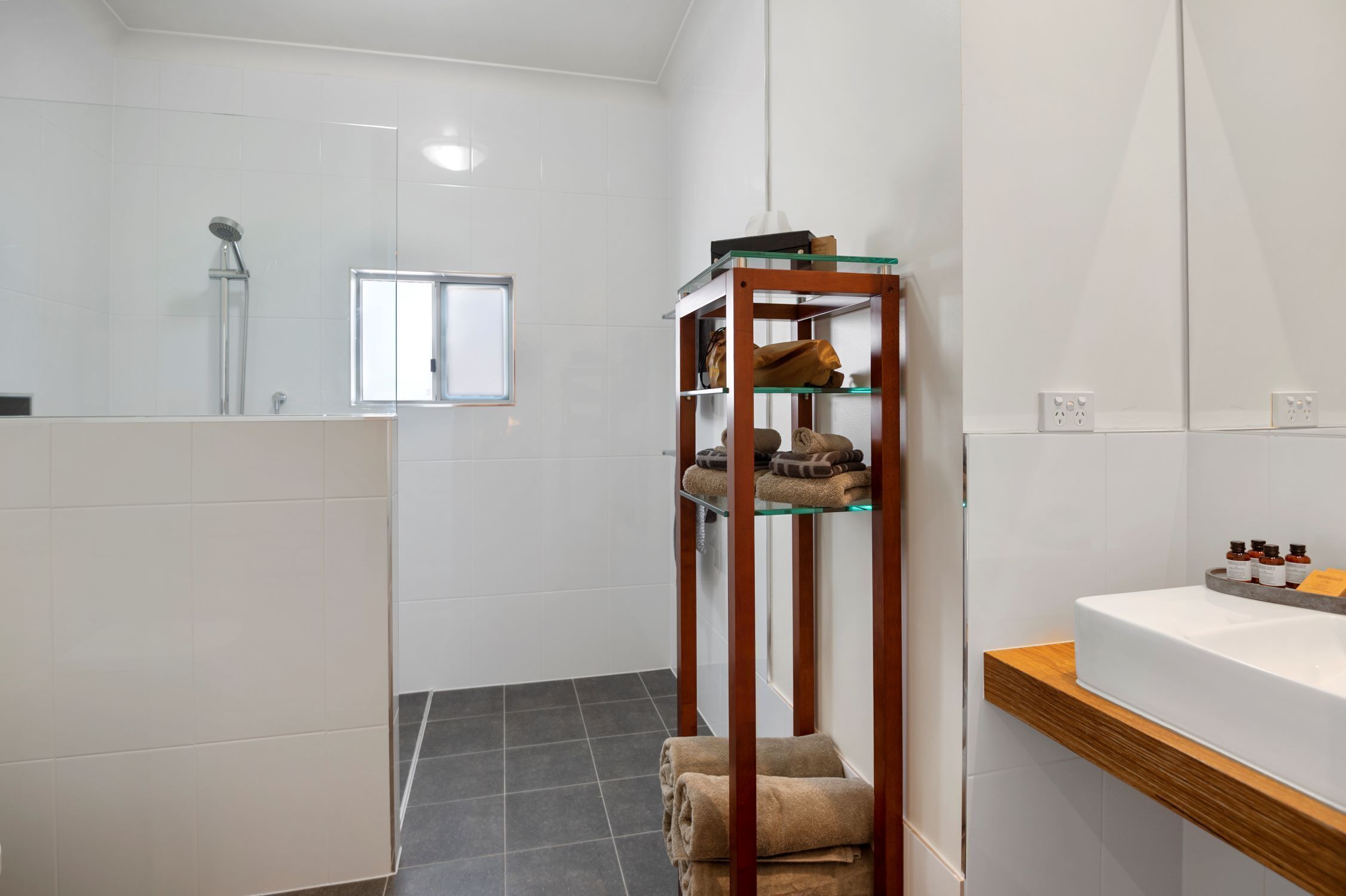 A modern bathroom with a glass shelf holding towels, a shower, and a vanity with a sink and toiletries.
