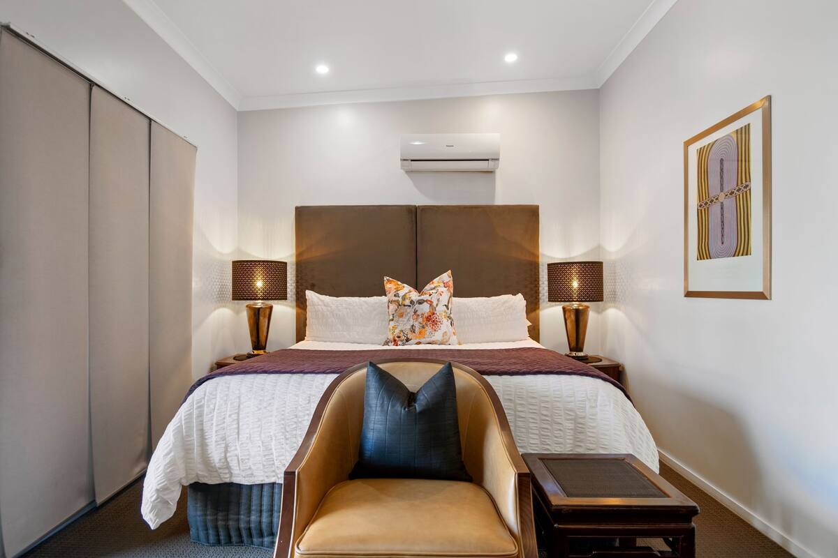 Habitación de hotel con una cama grande, dos lámparas de noche, una silla, una mesita y un equipo de aire acondicionado.
