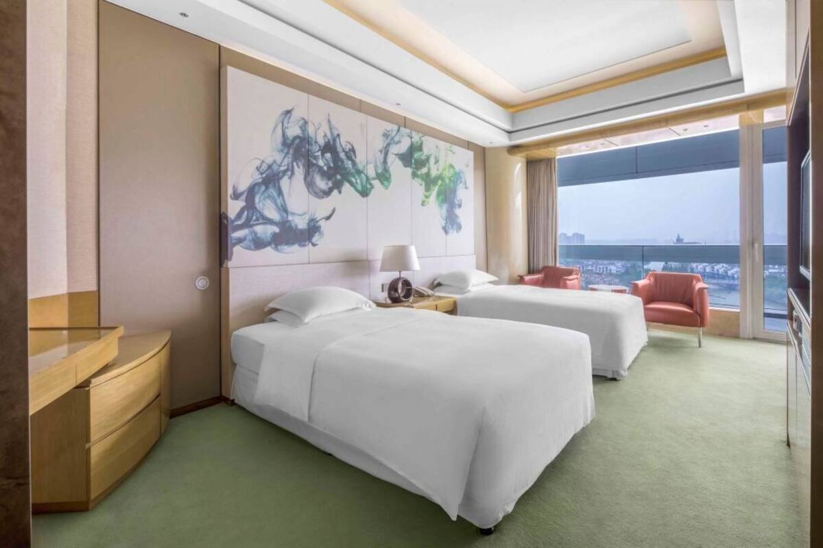 A hotel room with two beds, a desk, a chair, a large abstract painting, and a view of a cityscape.