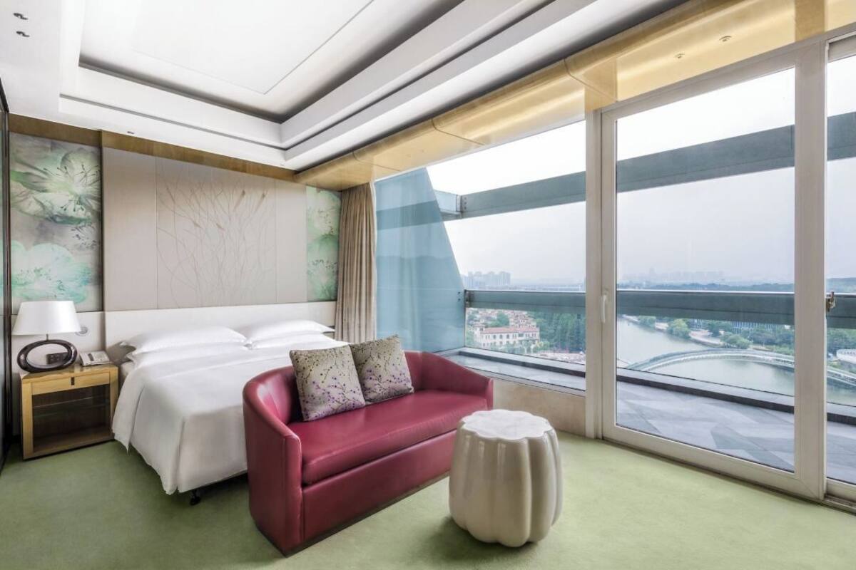 A hotel room with a bed, a red sofa, a nightstand with a lamp, and a large window with a view of a bridge and buildings.