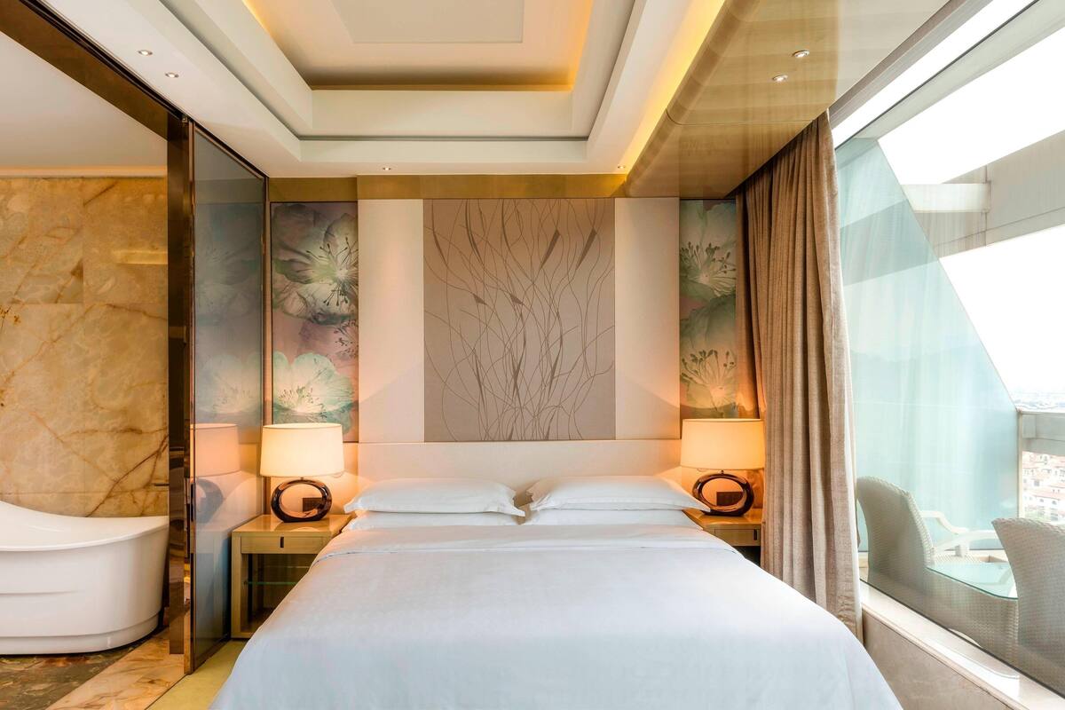 A modern hotel room with a large bed, a sitting area with a chair, a glass wall with a view, and a marble wall.