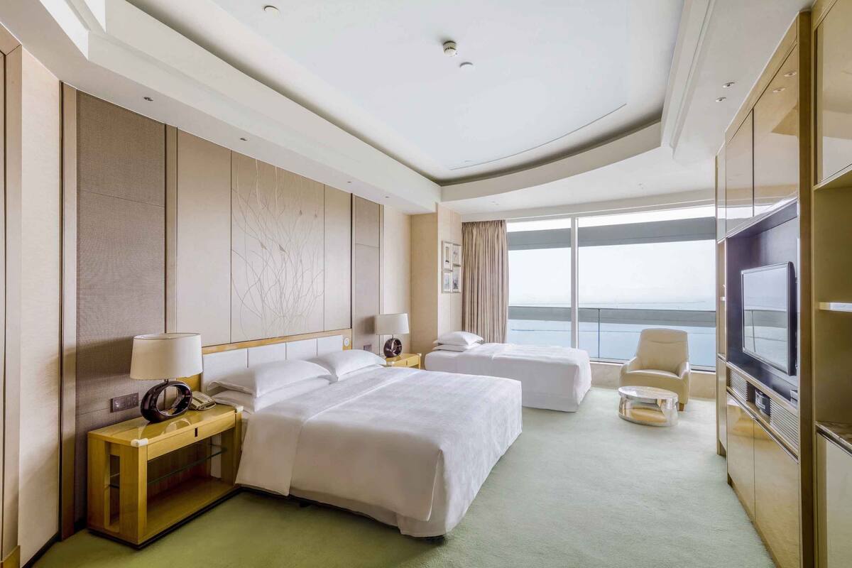 A hotel room with two beds, a TV, a desk, and a view of the sea.
