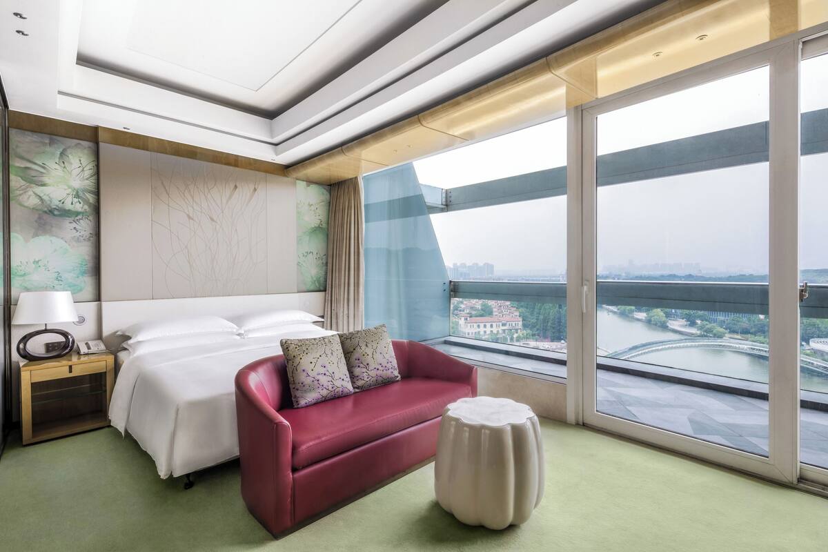 A hotel room with a bed, a red sofa, a small round table, a nightstand with a lamp, and a large window offering a view of a river and cityscape.
