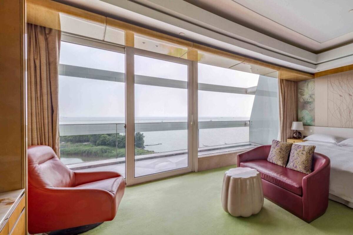 A hotel room with a large sliding glass door, a red sofa, a bed, and a small round ottoman.