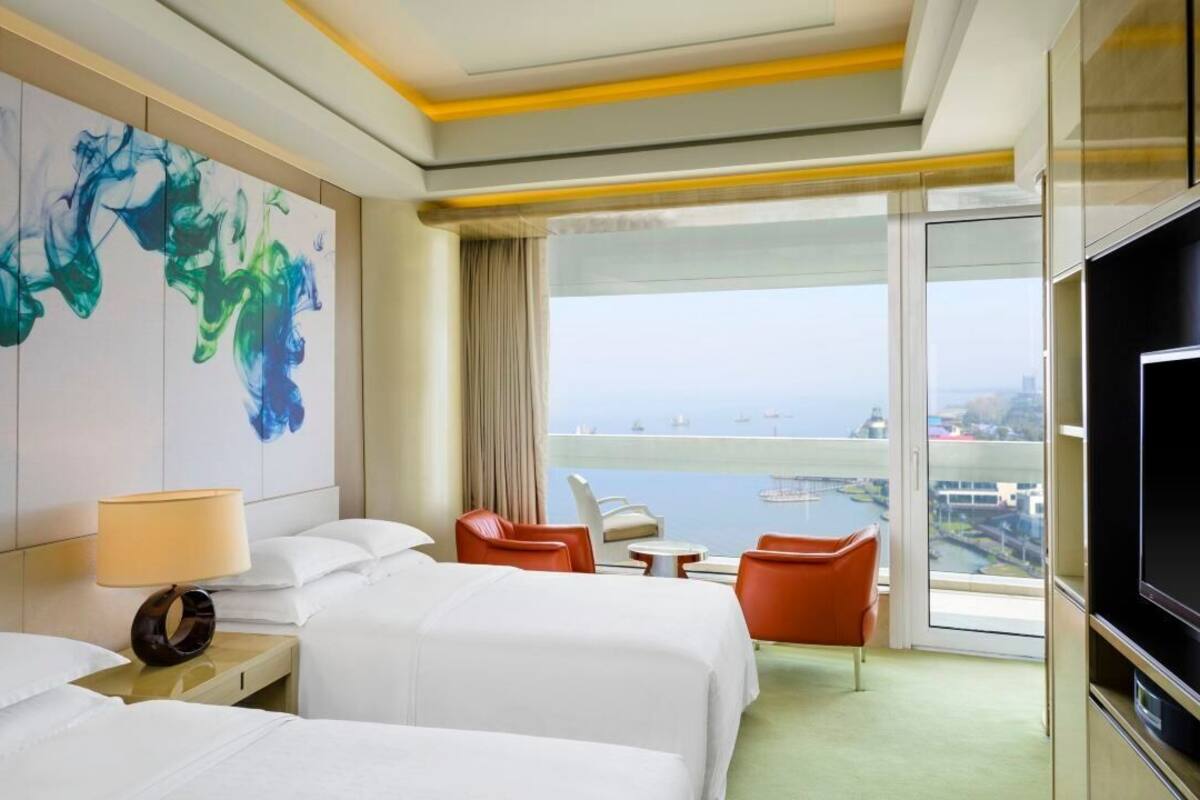 A hotel room with two beds, a TV, a painting, a balcony with a view, and a table with chairs.
