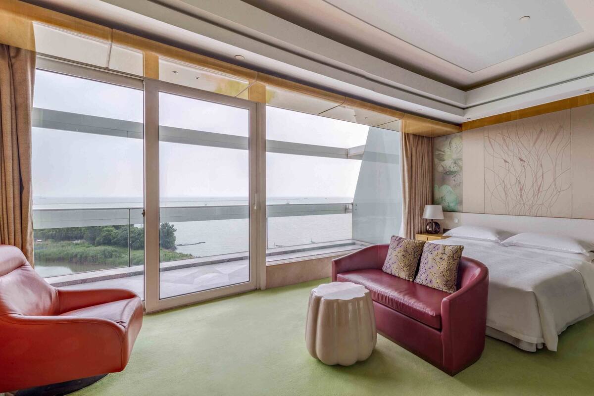 A hotel room with a large window, a bed, a red leather chair, a small ottoman, and a nightstand with a lamp.
