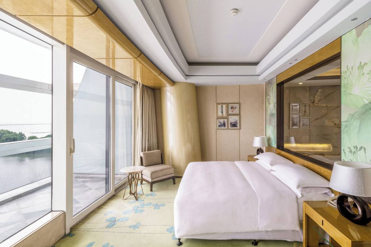 A hotel room with a large bed, a bedside table with a lamp, a chair, and a large window offering a view of a river and greenery.