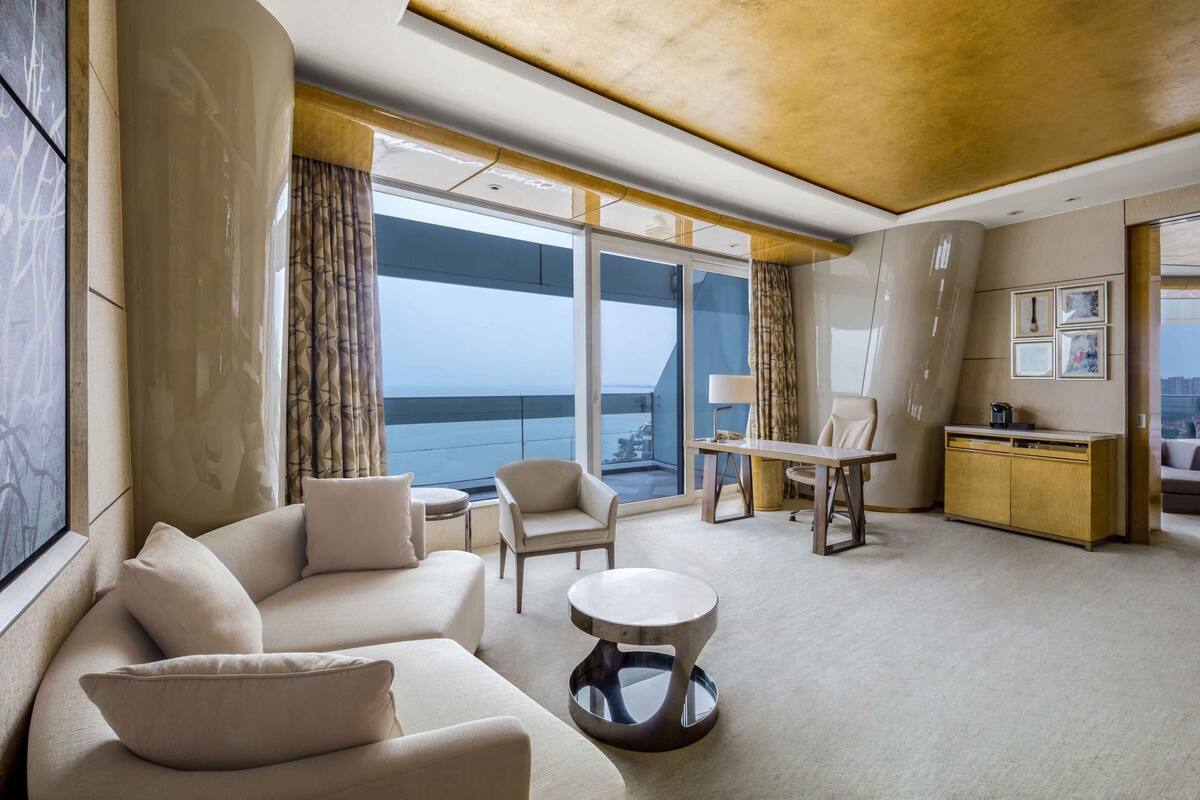A modern hotel room with a sofa, armchairs, a small table, and a view of the ocean.