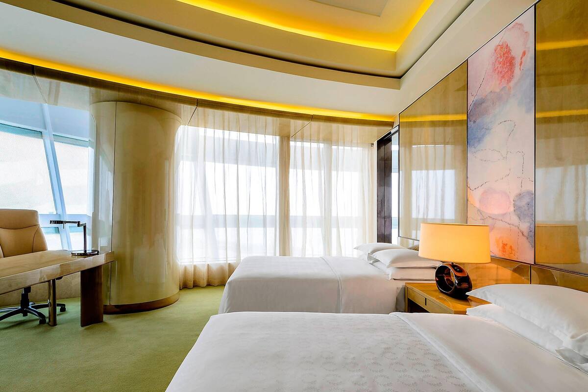 A hotel room with two beds, a desk, a chair, a lamp, and a large window with sheer curtains.