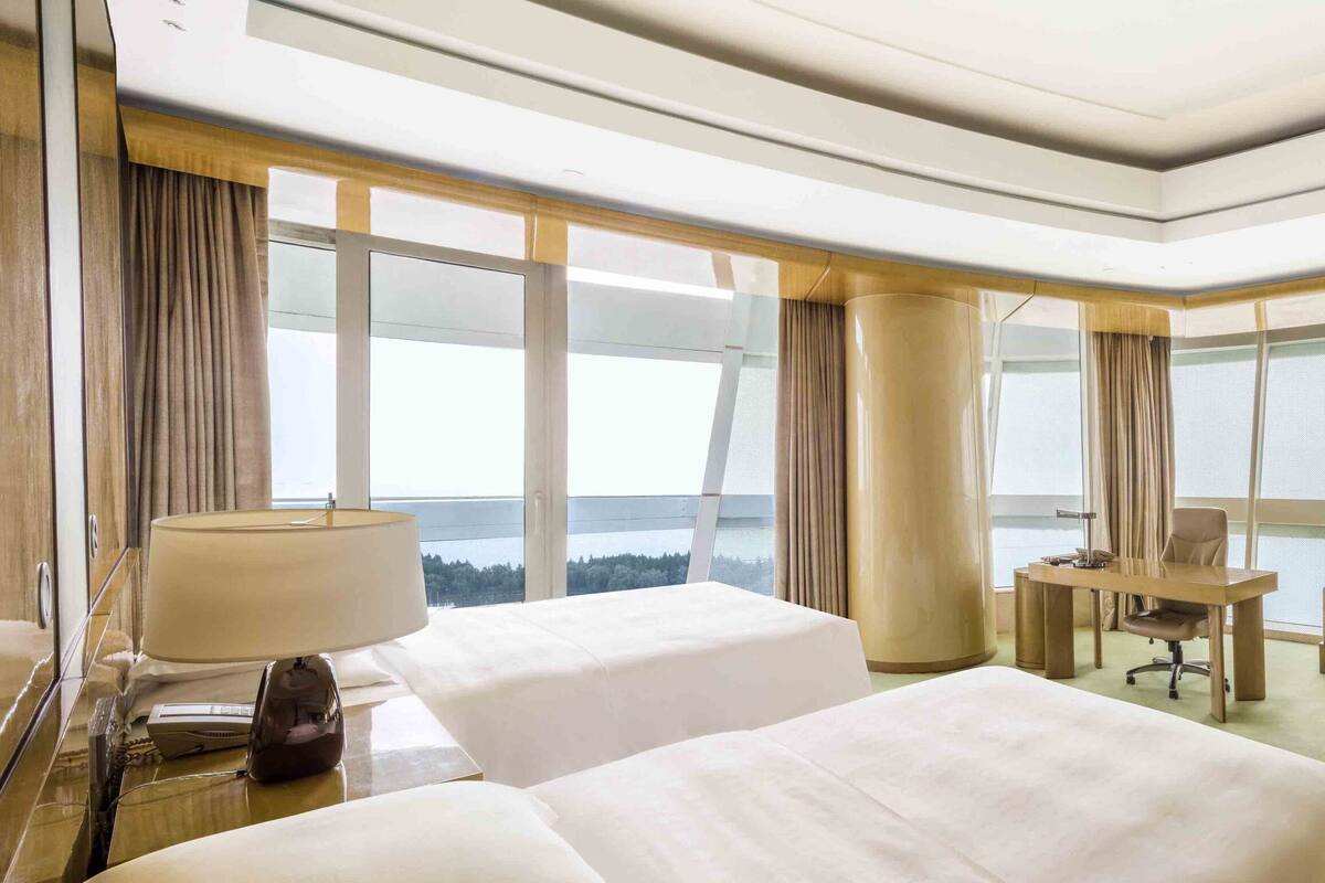 A hotel room with two beds, a desk, a sofa, a TV, and large windows with curtains.