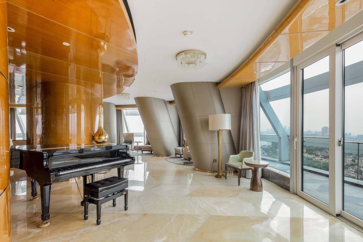 A spacious room with a grand piano, a chandelier, and a large window offering a city view.