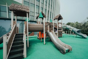 Un parc infantil amb tobogans i elements per escalar, amb un edifici de grans finestres al fons.