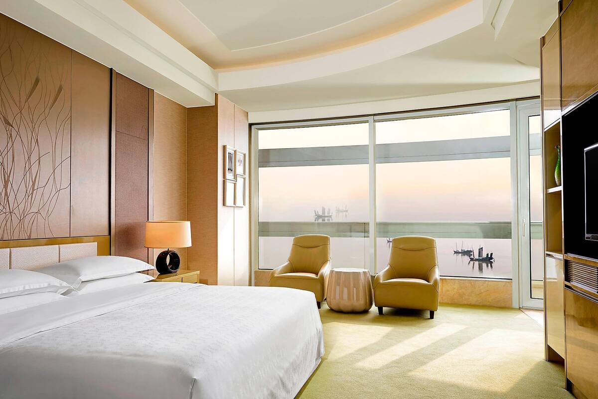 A hotel room with a large bed, two armchairs, a small table, a TV, and a large window with a view of a cityscape.