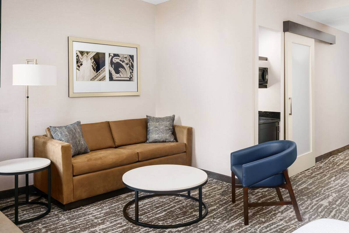 Une chambre d’hôtel avec un canapé marron, deux tables rondes, une chaise bleue, une œuvre d’art encadrée au mur et une porte donnant sur une cuisine.