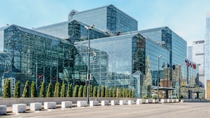 Een modern glazen gebouw met een geometrisch ontwerp, omgeven door een rij netjes bijgesnoeide bomen en witte plantenbakken.