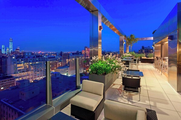 Una terraza en la azotea con mobiliario exterior, vistas de la ciudad y una jardinera grande con flores.