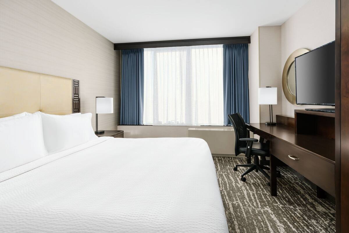 Une chambre d’hôtel avec un grand lit, un bureau, une chaise, une télévision et une fenêtre avec des rideaux.