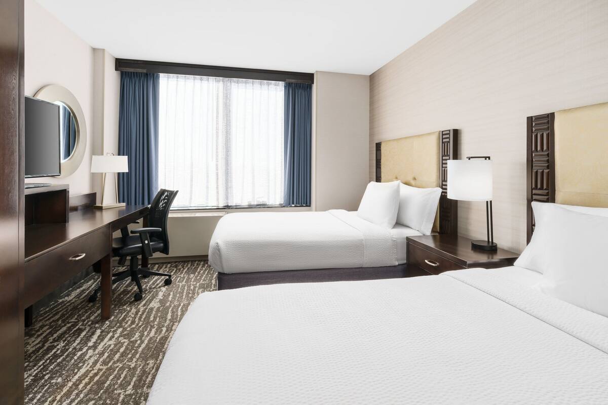 Une chambre d’hôtel avec deux lits, un bureau, une chaise, une télévision et une fenêtre avec des rideaux.