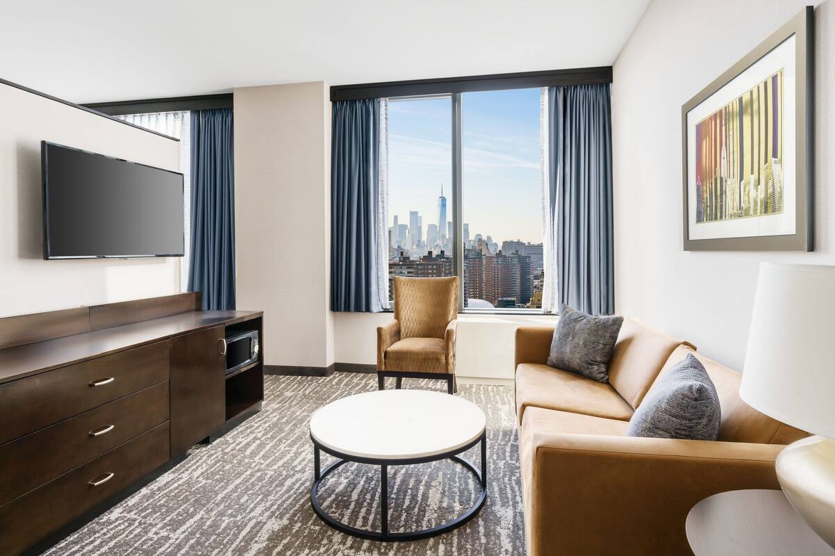 Une chambre d’hôtel moderne dotée d’une grande fenêtre donnant sur la ville, d’un téléviseur à écran plat, d’un canapé, d’un fauteuil et d’une table basse.