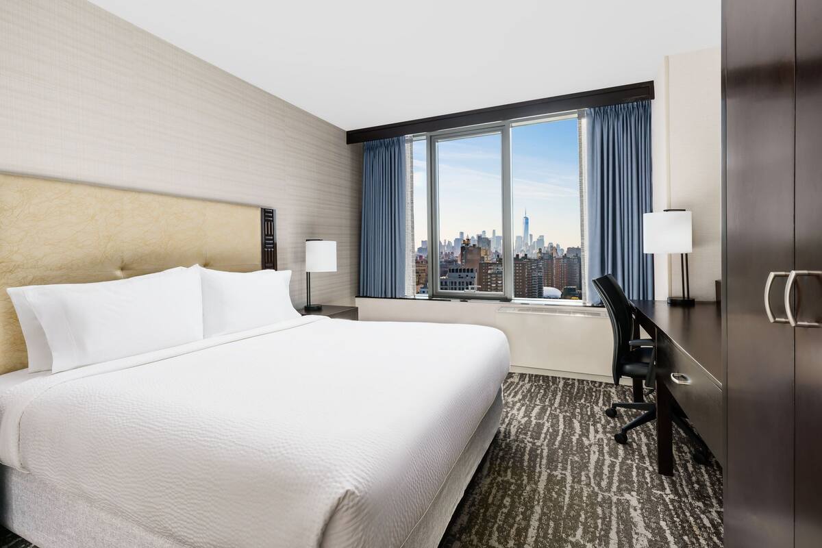Une chambre d’hôtel avec un grand lit, un bureau et une vue sur le paysage urbain, visible à travers une grande fenêtre.