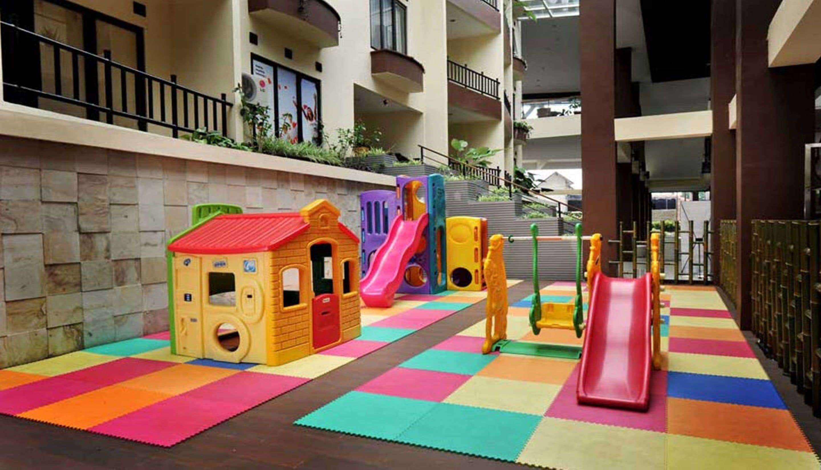A children's play area with colorful play structures and mats, located in a multi-story building with balconies and a view of the surrounding area.