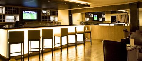 A modern bar area with a long counter, multiple bar stools, and two televisions mounted on the wall.