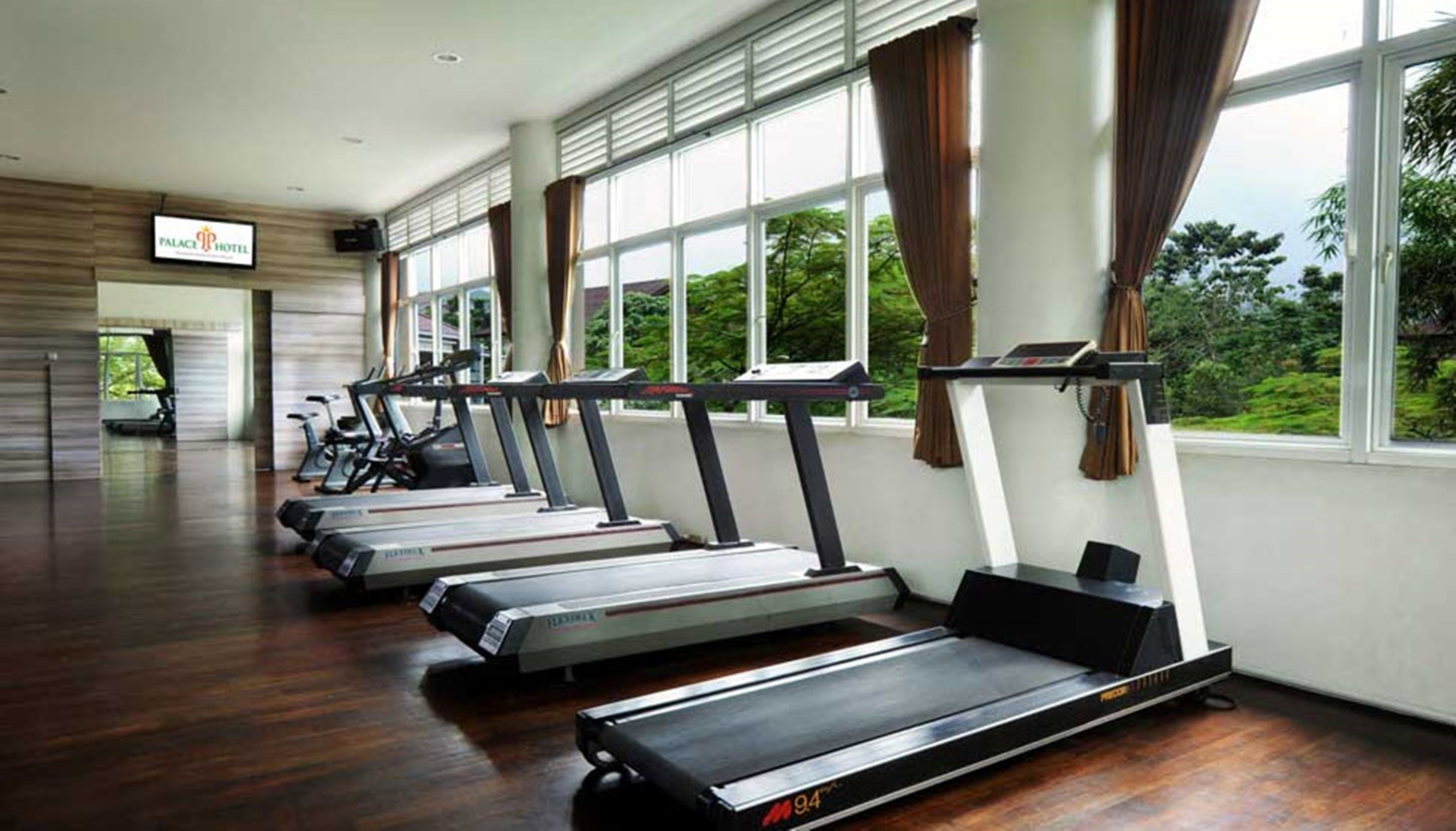 Ein Fitnessraum mit Laufbändern, die an einer Wand mit großen Fenstern angeordnet sind, einem Schild für das Hotel und einer Tür zum Ausgang.